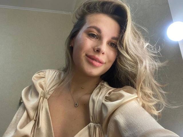 MoonriseGoddessx-hot with blonde hair, pink lips, wearing a beige, puffy-sleeve top, and a small necklace, smiling softly at the camera. Bright light in the upper right.