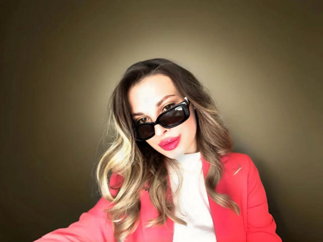 MoonriseGoddessx-hot with blonde ombre hair, wearing black sunglasses, red blazer, white turtleneck, and bright pink lipstick, smiling slightly, against a dark greenish-brown background.