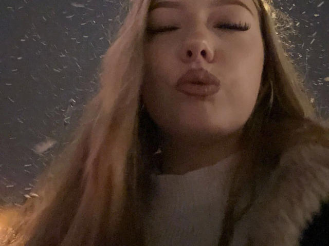 Sexy-naty-hot with pouty lips, long blonde hair, and closed eyes, blowing snow in a dark, cold night. She's wearing a fur-lined jacket, face slightly blurred.
