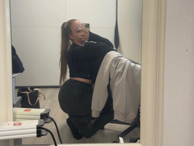 Sexy-naty-hot with a big ass, wearing a black crop top and dark jeans, taking a mirror selfie. Long brown ponytail, smiling. Mirror reflection, gray jacket on chair.