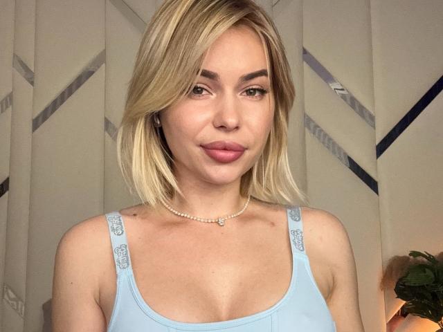 AmelyDragons a les cheveux blonds, la peau claire, et porte un débardeur bleu clair avec des bretelles sur lesquelles est écrit "SAC", ainsi qu'un collier de perles. Elle sourit, et ses lèvres sont roses..