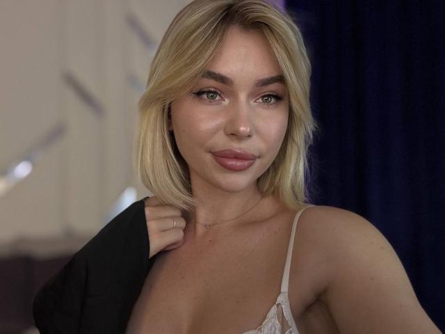 AmelyDragons met blond haar, groene ogen en volle lippen, die een kantachtig wit beha draagt, een zwarte kledingstuk vasthoudt, zachtjes glimlacht en haar borsten en subtiele decolleté laat zien..
