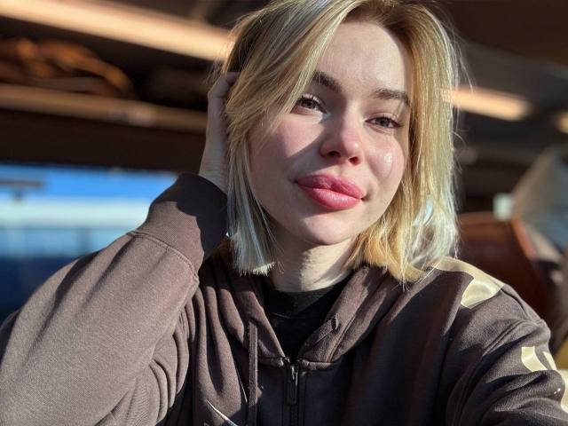 Amely: Dragões com cabelo loiro, lábios rosados e moletom marrom, segurando a cabeça, luz do sol no rosto, pele levemente brilhante, selfie casual, sorriso suave, fofa, com uma vibe relaxada..