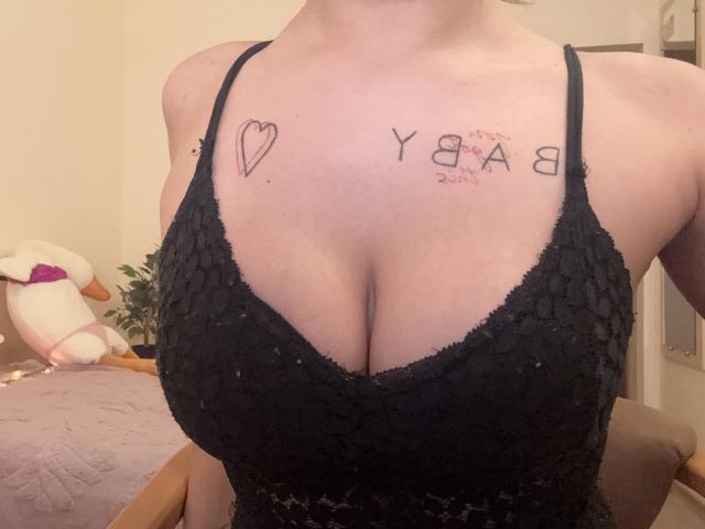 Os grandes seios de Kametia em um vestido preto de renda, com tatuagens acima do seu colo, com um coração e "Y8A8" em sua pele clara, mostrando um pouco do seu pescoço e peito..