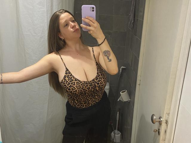 BeluSexy69 with large tits in a leopard print top, black skirt, holding a phone mirror selfie, showing a tattoo on her right arm, in a bathroom.