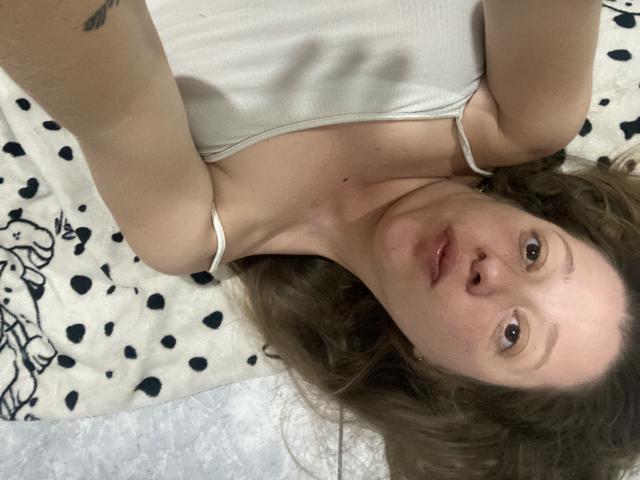 BeluSexy69 with wavy brown hair, white tank top, and a tattoo on her left arm, lying on a spotted blanket, looking up at the camera with a neutral expression.