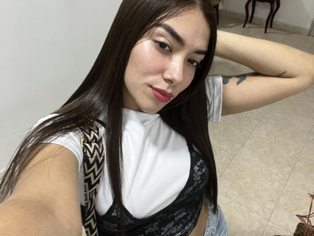 SharonKlart with long brown hair, wearing a white tee and black lace bralette, showing her tits, nose ring, and tattoo, taking a selfie with one arm raised.