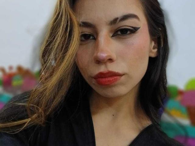 KatherinJohns with serious face, dark makeup, bold eyeliner, red lipstick, black top, long brown hair with blonde highlights, soft skin, slightly parted lips, blurry colorful background.