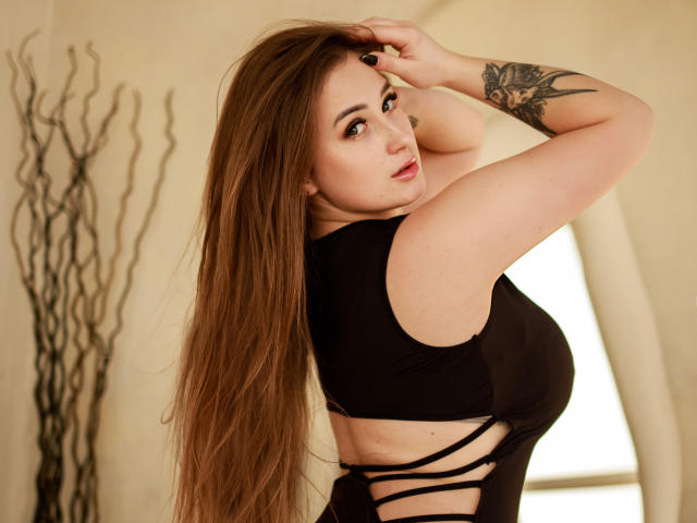 GentleAdellee mit langen, braunen Haaren, schwarzem Top ohne Ärmel, das den Rücken freilegt, Tätowierung am rechten Arm, der rechte Arm ist angehoben, sie schaut über die Schulter, helle Haut, roter Lippenstift, sanftes Lächeln, Schlafzimmerumgebung..