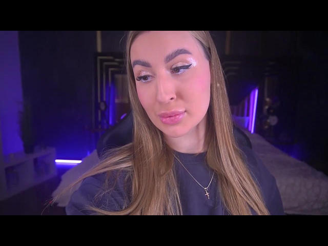 LiaBrazzerr has long brown hair, pink lipstick, and a cross necklace, looking down with slightly parted pink lips, wearing a black shirt, in a purple-lit room.