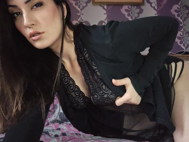 Alejandra Delaney mit langem, dunklem Haar, einem schwarzen Spitzen-BH und einem transparenten schwarzen Oberteil, die nach vorne gebeugt ist, eine Hand auf der Hüfte, wobei ihre Brüste und schwarze, schmale Unterwäsche zu sehen sind..