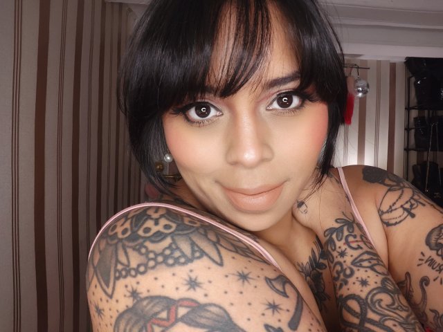 Chili, a sexy Asian woman with black hair and bangs, is topless, showing off her heavily tattooed shoulders and breasts, smiling seductively at the camera.