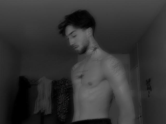 Luccamore is a shirtless dude with tattoos, dark messy hair, and a beard, looking down, showing a slim, toned upper body and a cross tattoo on his chest.
