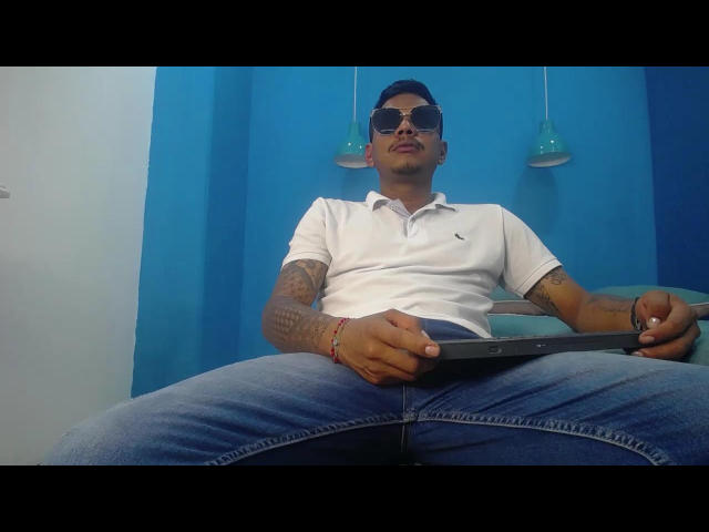 SamSexy69, con tatuaggi, indossa una polo bianca, jeans blu e occhiali da sole, seduto con le gambe divaricate, tiene in mano un tablet, con un'espressione seria, in una stanza dai colori vivaci..