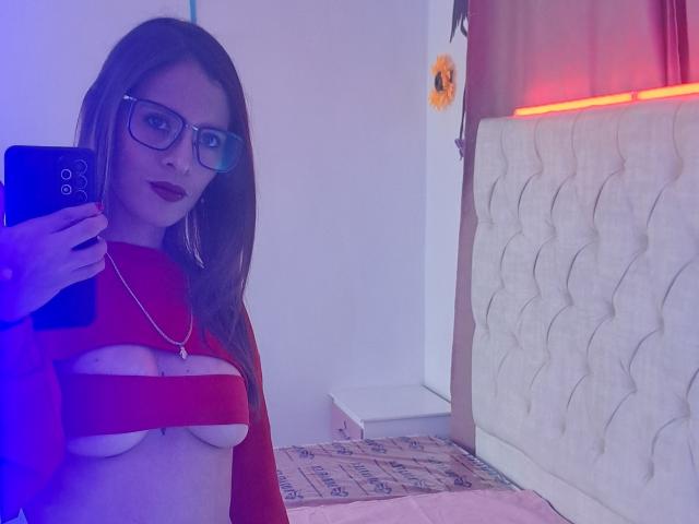 AnnyQueen with glasses, red top, exposing her tits, taking a selfie in a bedroom with a tufted headboard, holding a blue phone, and wearing dark lipstick.