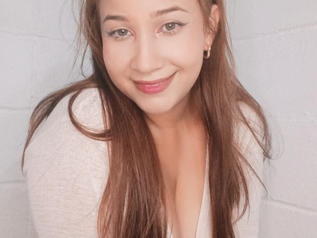 AllannaHanks with long brown hair, light skin, and small gold earrings, smiling at the camera, wearing a white top with a low neckline, showing her tits.