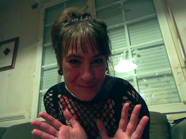 AcideCyprine with brown hair in a messy bun, wearing a black fishnet top, hands up, smiling, has a nose piercing and ear gauges, sitting indoors.