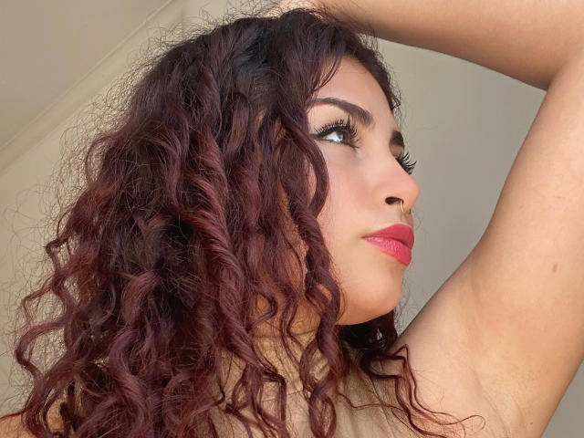 SweetSolei has curly, dark red hair, brown eyes, and pink lipstick. She's raising her arm, showing a nose ring and looking to the side. Her tits are not visible.