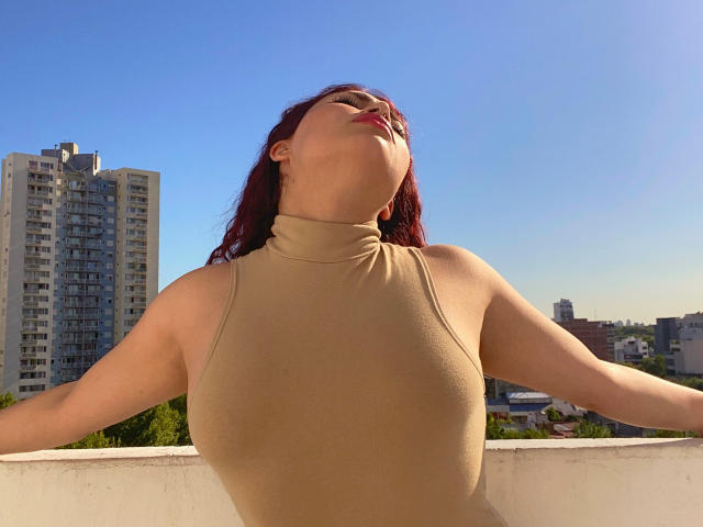 SweetSolei with red hair, eyes closed, head tilted back, wearing a beige turtleneck, arms spread, sunlight on her face, standing on a rooftop, clear blue sky.