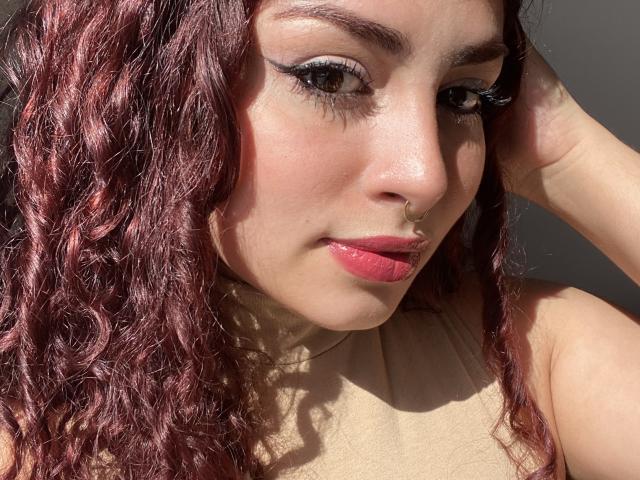 SweetSolei's face, showing pink lipstick, curly red hair, nose ring, black eyeliner, and sunlit skin, with a hand touching her head. Boobs not visible.
