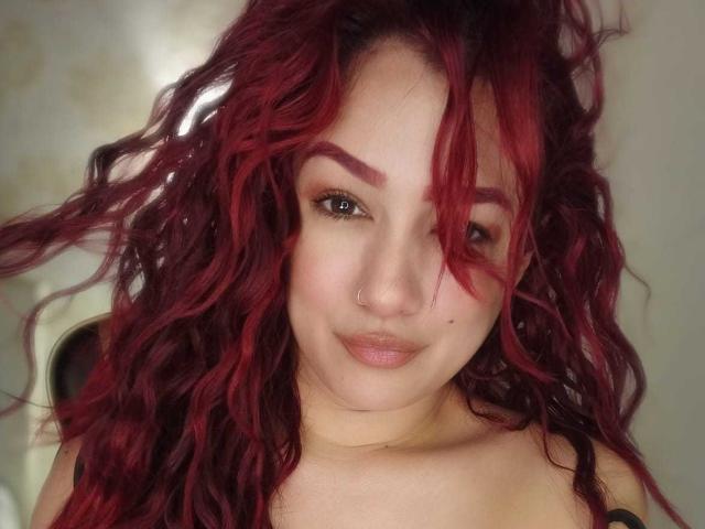 SammyJohnson has curly red hair, a nose piercing, and a slight smile. She's looking at the camera with her tits barely visible in the frame.