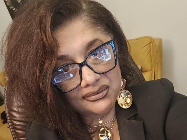 ClassybutNaughty with brown hair, blue glasses, and dark lipstick, wearing a black blazer and gold earrings, looking slightly confused. Boobs not visible, focused on face.