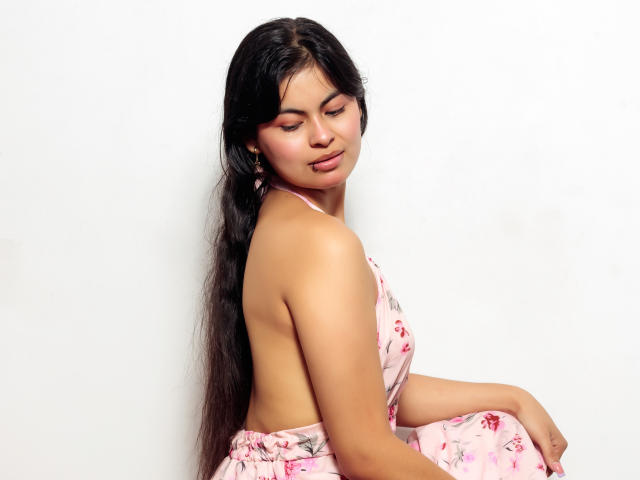 CrystallThonpsom with long black hair, pink floral dress, and bare back, sitting, eyes closed, soft smile, showing her small tits and side-ass. White background.