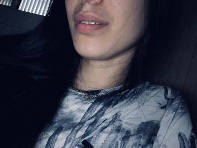 DianaHottie's face, showing pink lips, small white teeth, and a bit of a chin, with black hair, wearing a gray and black tie-dye shirt, and a gold necklace.