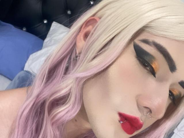 Tokyo Evans, con cabello degradado de rubio a rosa, delineador de ojos negro grueso, lápiz labial rojo, un piercing en la nariz y un brazo tatuado, está acostada sobre un cabecero negro acolchado. No se ven los senos..