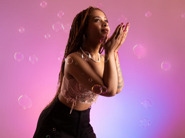 KendallQuinn with brown braids, pink lace top, black pants, hands up, bubbles around, pink to purple gradient background, smiling, tattoos on arms, pink nails, soft lighting.