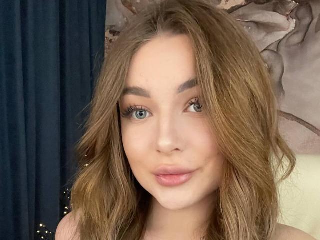  AlexandraFlirty has blue eyes, wavy brown hair, and full pink lips. She's smiling softly with natural makeup. Her tits are partially visible in the top frame.