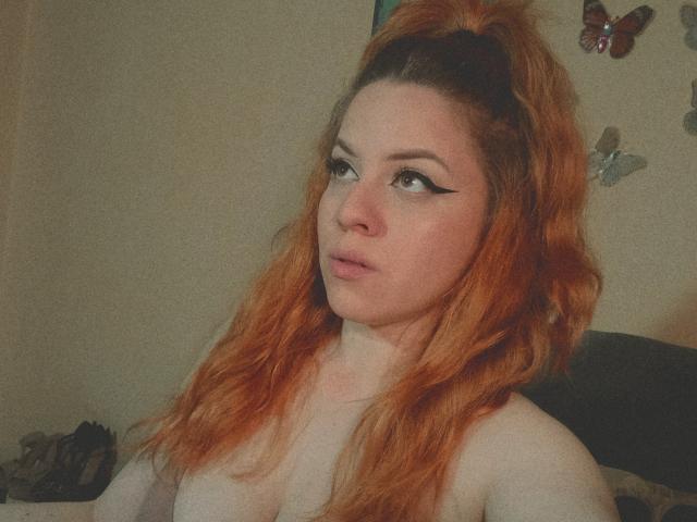 SpicyGrace, con i capelli rossi e ondulati raccolti in una coda alta, con l'eyeliner nero, guarda di lato con un leggero sbuffo, senza top, mostrando il seno. Decorazioni murali a forma di farfalla..