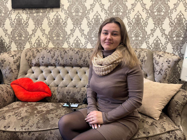KarlaKushh, blonde, wearing brown turtleneck, scarf, and black tights, sits on a beige floral sofa with red lips pillow, remote, and beige pillow. Her tits are small.