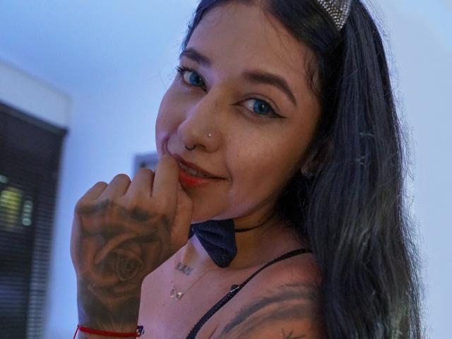 EmmaRachel with blue eyes, black hair, and tattoos on her hand and arm, biting her finger, wearing a black bow tie, and red wristbands, smiling slightly.