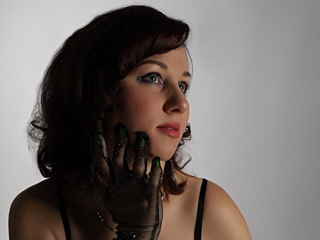 MirandaBrooks has dark, wavy hair, green eyeshadow, and is looking to the side with a slight smile. She's wearing a black mesh glove with green nail polish.