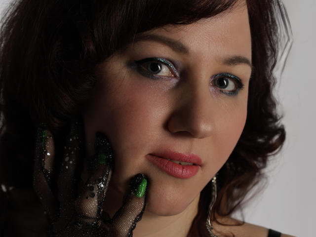 MirandaBrooks has green glitter on her black lace gloves, with blue eyeshadow and pink lipstick, touching her cheek with her left hand. She's looking at the camera.