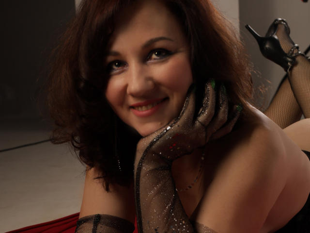 MirandaBrooks with curly brown hair, smiling, wearing sheer black gloves, and black high heels, resting her chin on her hand, showing off her tits, and green-painted nails.