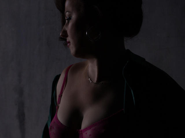 MirandaBrooks with dark hair, wearing a pink lace bra, showing her tits, in a green jacket, side profile, dark shadow, subtle necklace, earrings, dim light.