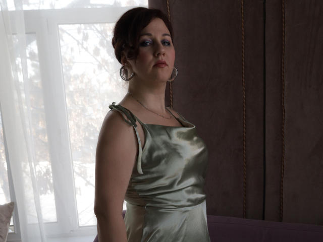 MirandaBrooks with pale skin, curly brown hair, blue eyeshadow, wearing a shiny green satin dress, silver hoop earrings, and a necklace, standing in a dimly lit room.