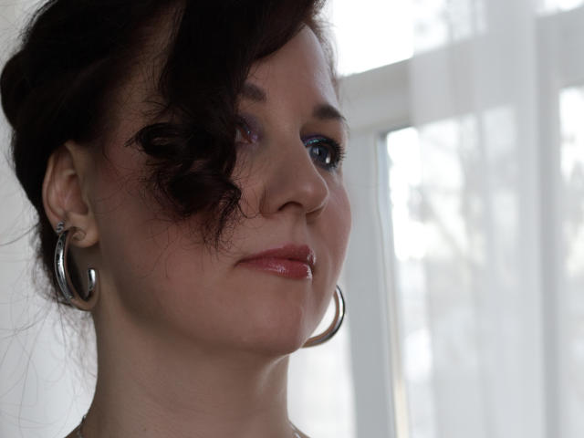 MirandaBrooks with dark curly hair, blue eyeshadow, pink lips, and silver hoop earrings, looking to the side with a slight smile. Soft light from the window.
