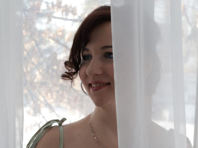 MirandaBrooks with curly red hair, blue eyes, and a silver necklace, peeks through a white curtain, smiling softly, showing her cute face and bare shoulders.