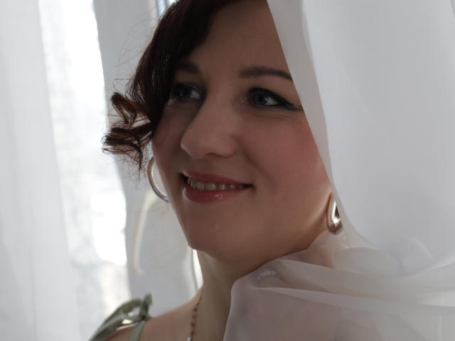 MirandaBrooks has fair skin, red curly hair, blue eyes, and a cute smile. She's wearing a sheer white fabric and silver hoop earrings, looking up with a gentle expression.
