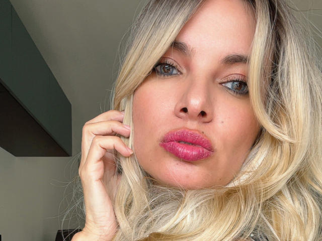SaraLoveX69 with blonde hair, blue eyes, pink lipstick, and a pouty face, touching her hair with her right hand. Her tits are not visible.