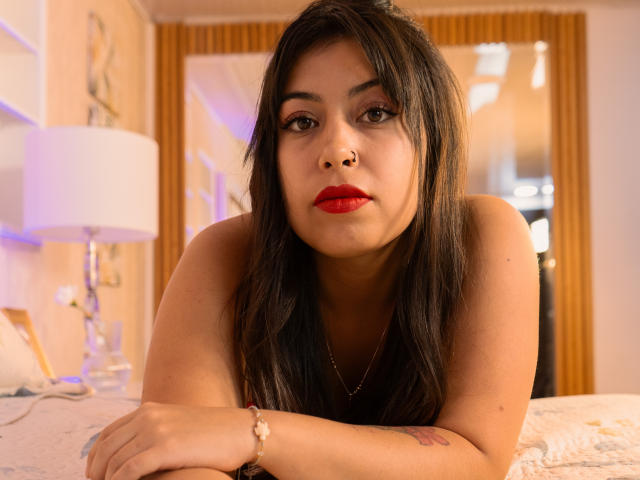ConnieSylvie, with long black hair, red lipstick, and a nose ring, leans on a bed, showing a tattoo on her right arm and a bracelet on her wrist.
