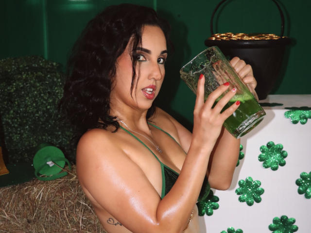 DianaClarke with curly black hair, green lingerie, holding a green glass, looking at it with a slight grin, showing her shiny tits and tattoo on her right arm.