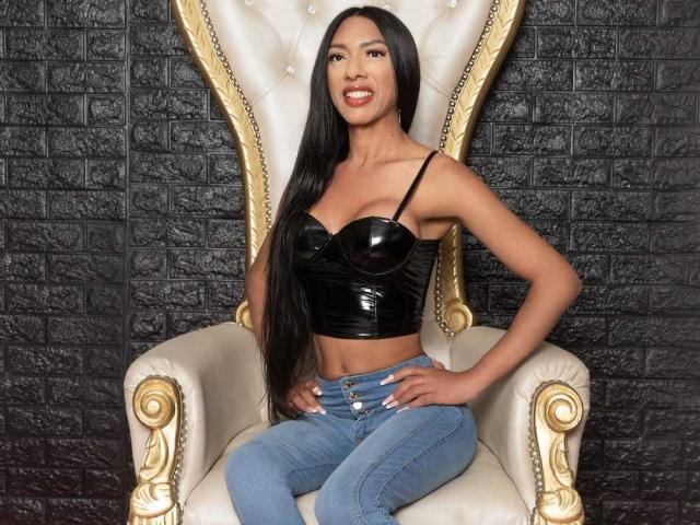 BrendaBigCock with long black hair, wearing a shiny black bustier and blue jeans, sitting on a gold-trimmed, high-back chair, hands on hips, smiling.