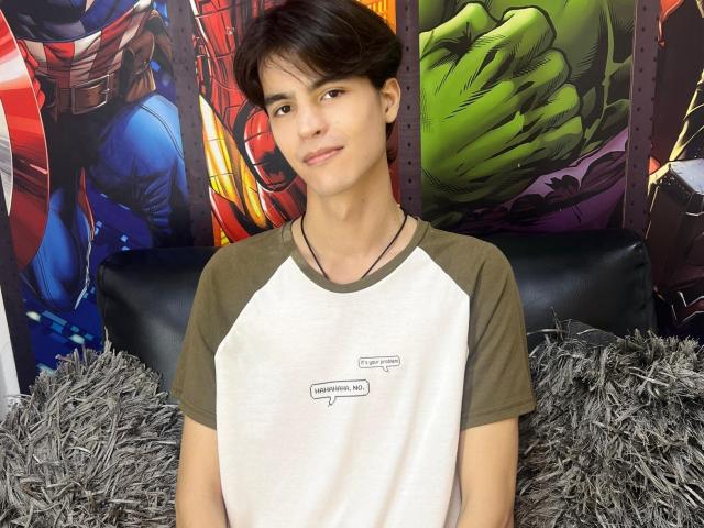 TylerMulber, young dude with brown hair, wearing a raggedy white and brown tee, smiling, black necklace, sitting on fuzzy gray pillows, Marvel comic posters in background.