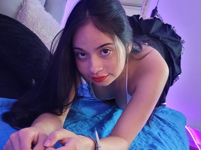ZuriLaoni, com cabelos pretos longos, batom vermelho e uma camiseta branca, apoiada em uma cama azul, olhando para a câmera com um leve sorriso, vestindo um vestido preto com amarração..