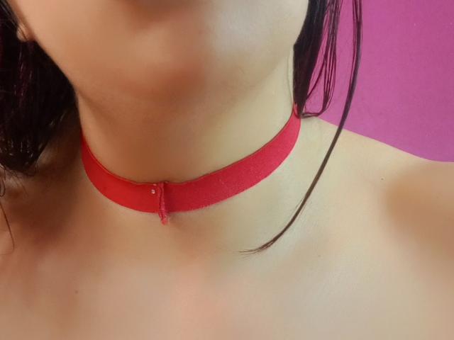 SublimeMature's neck with a red choker, skin is light, black hair in the frame, upper chest exposed, background is pink. Boobs not visible, focus on choker and neck.