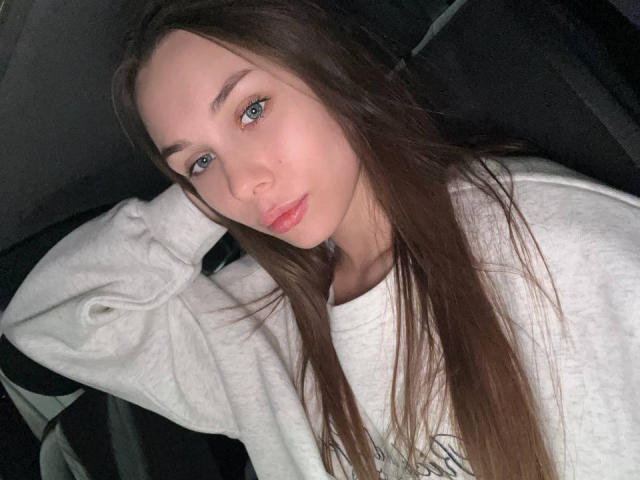 ElsaSexxy with long brown hair, blue eyes, and pink lips, wearing a white shirt, lying down with a relaxed, slightly seductive expression, looking at the camera.